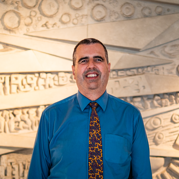 Head and shoulders portrait of Ben, wearing a blue shirt and multi-coloured tie, standing in front of a textured light-coloured background.