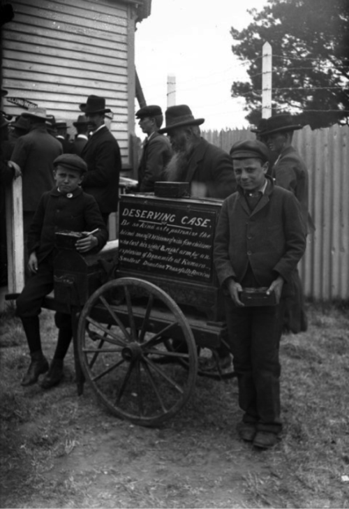 Black-and-white historical photo of 2 children holding bowls beside a handcart with a sign reading “Deserving Case”, with adults gathered behind.