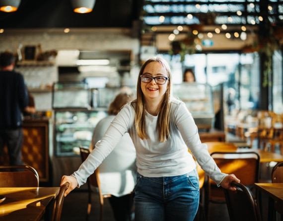 Young woman with glasses standing between tables and chairs in a café.