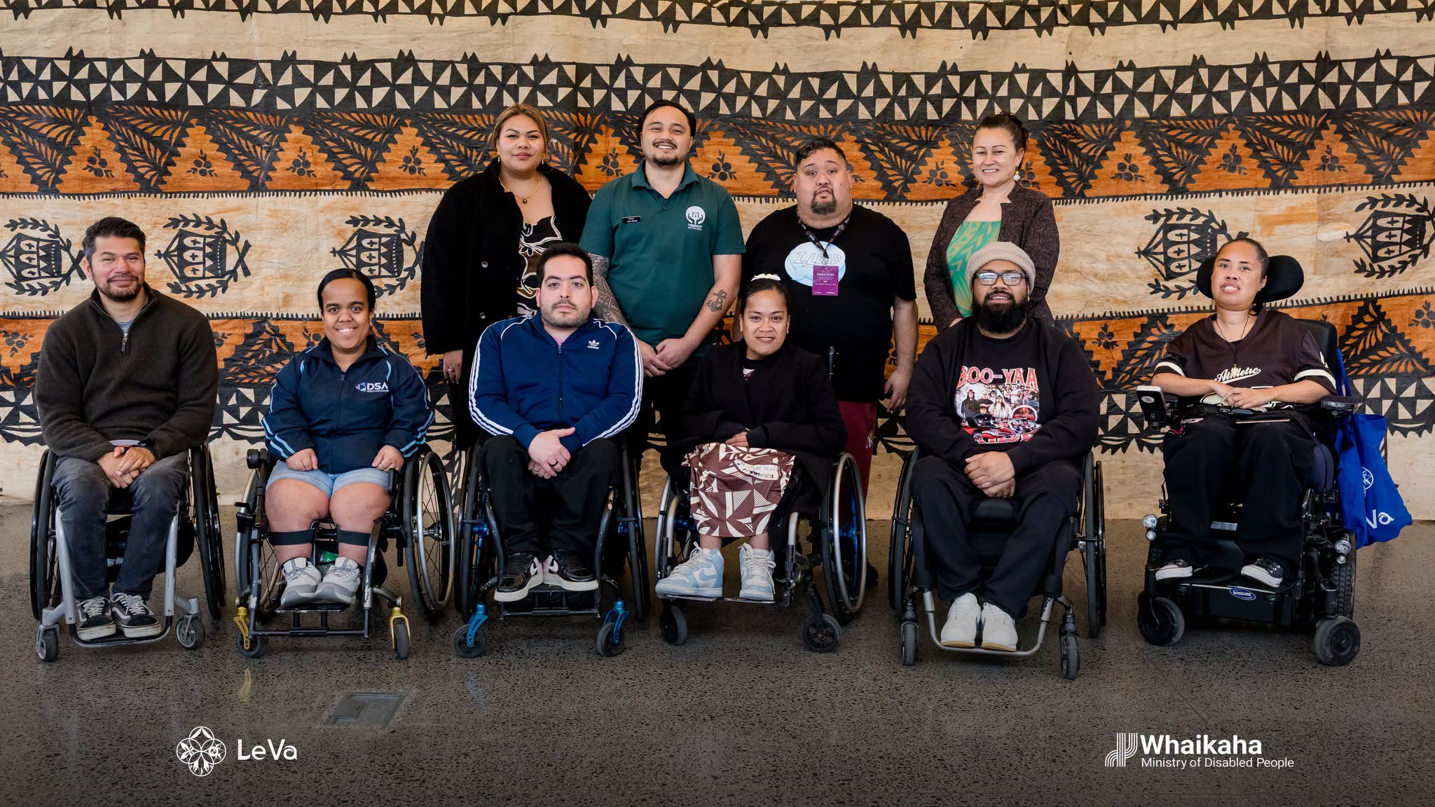 10 people in a group photo of 2 rows. In the front row are 6 people in either wheel or power chairs.