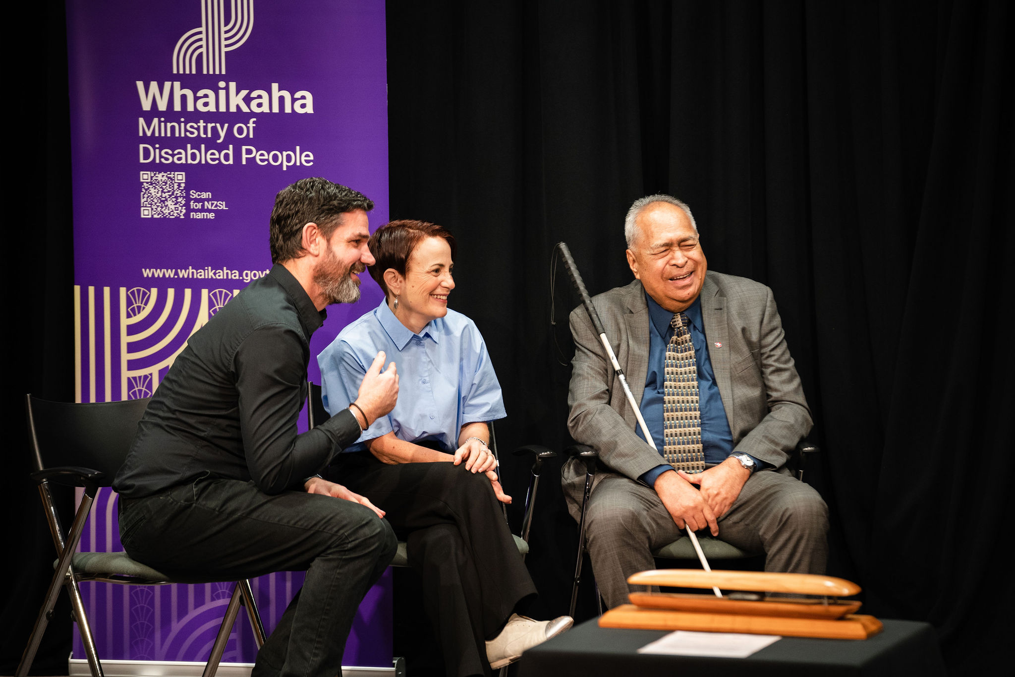 NZSL interpreter, Paula Tesoriero & Maaka Tibble seated on a stage. They are smiling and laughing together. A purple Whaikaha banner is behind them and Maaka is in a suit and holding a long silver and black cane