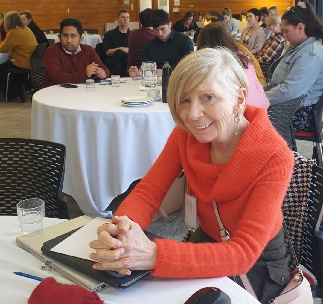 Pauline, a woman with white hair and a red jersey, sits at a table in a conference room and smiles at the camera