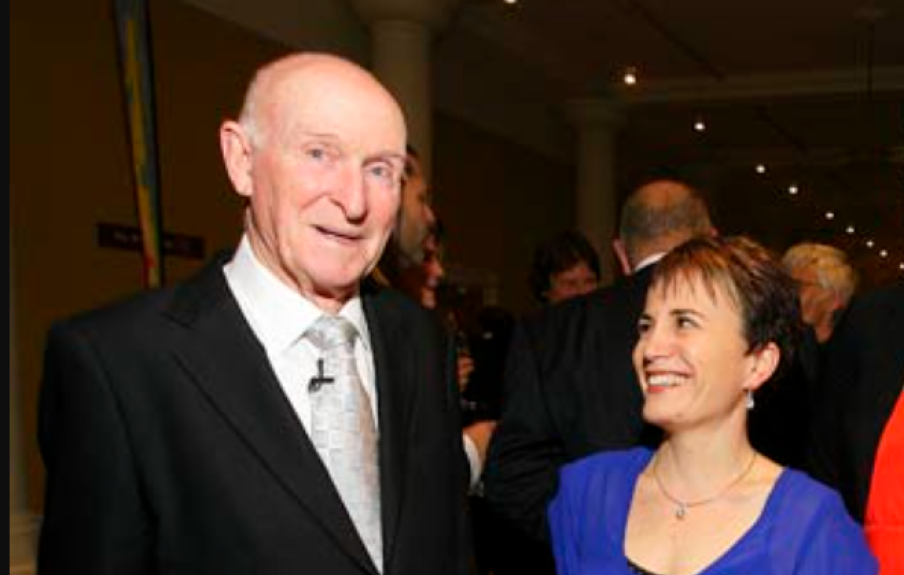 Sir Murray Halberg smiling at the camera in a black suit with a silver tie, Chief Executive Paula Tesoriero is standing to his right in a blue dress smiling up at him.