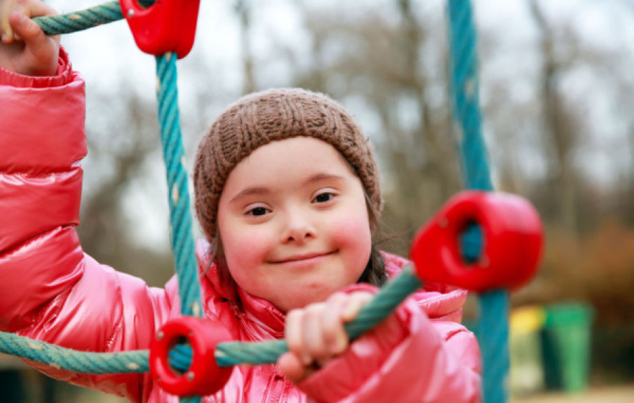 A young girl with Down syndrome smiles as she climbs a structure in a playground